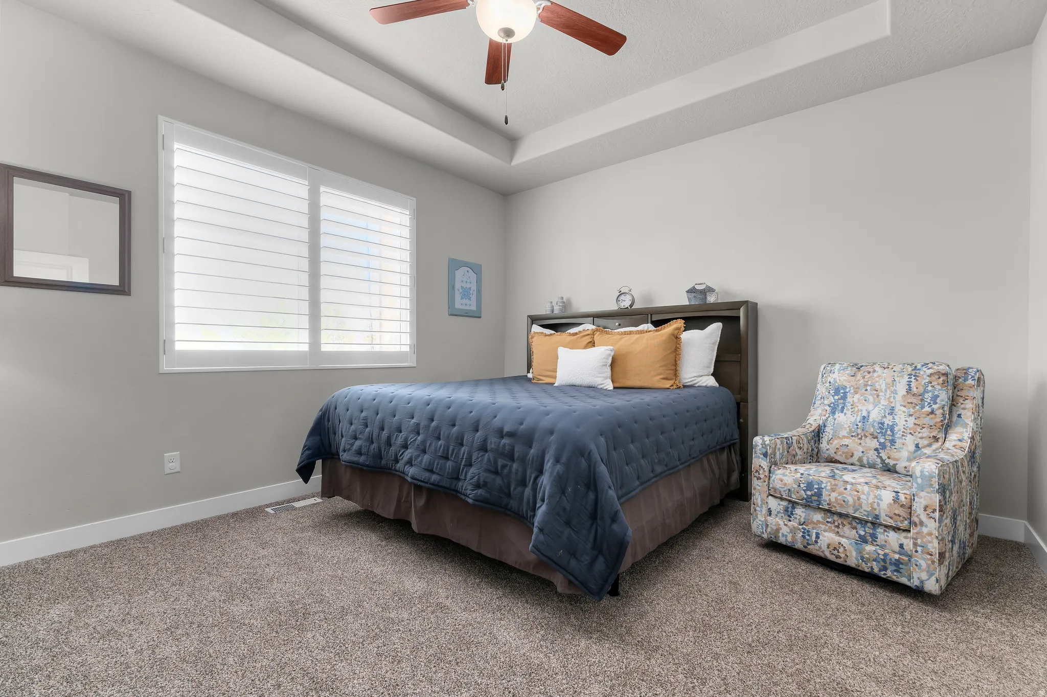 Bedroom featuring a tray ceiling with a ceiling fan, neutral carpet flooring, and a window with plantation shutters