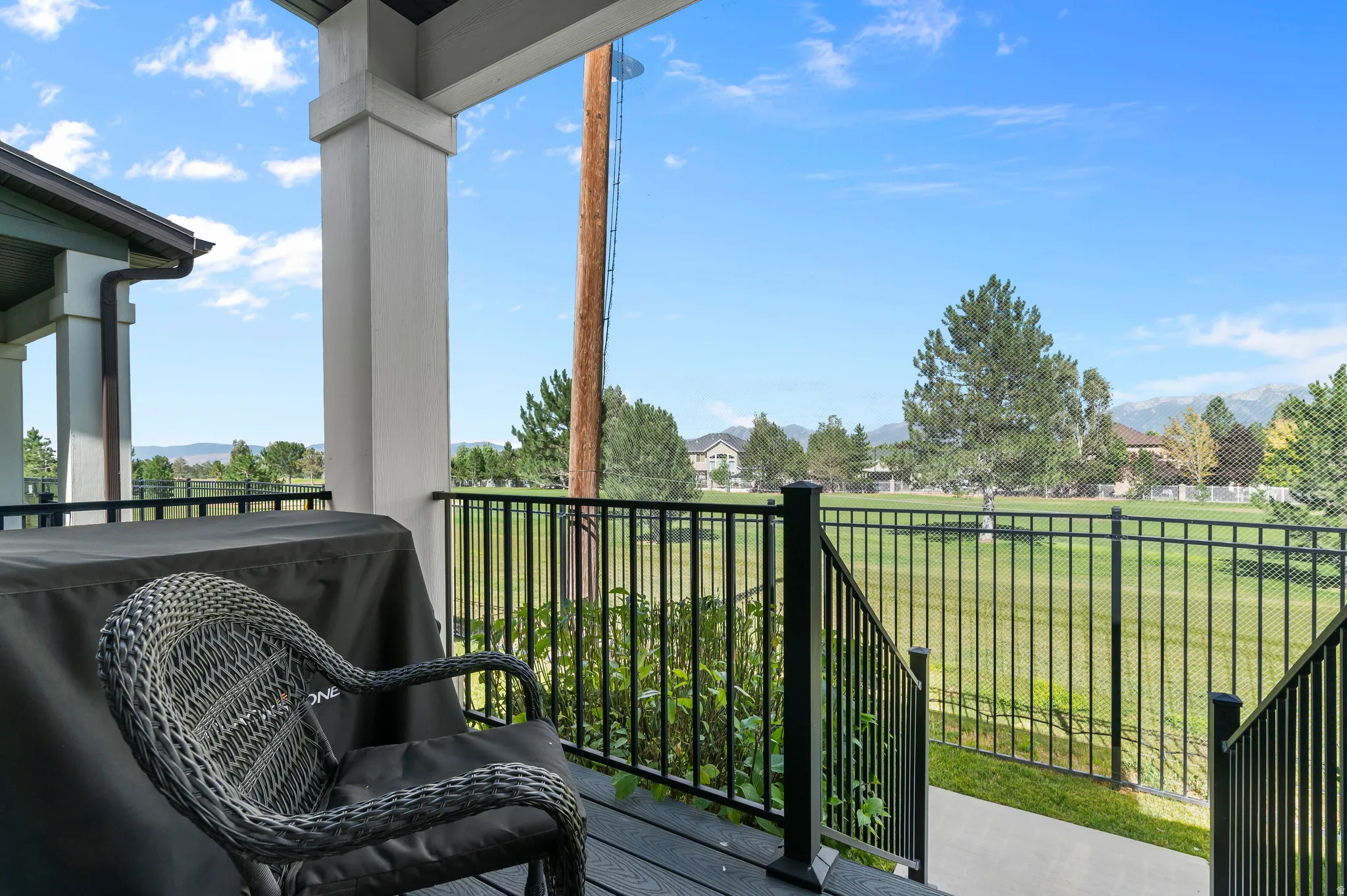 HOME EXTERIOR - REAR Covered outdoor deck with composite decking, white structural pillars, and black metal railing overlooking a landscaped green space