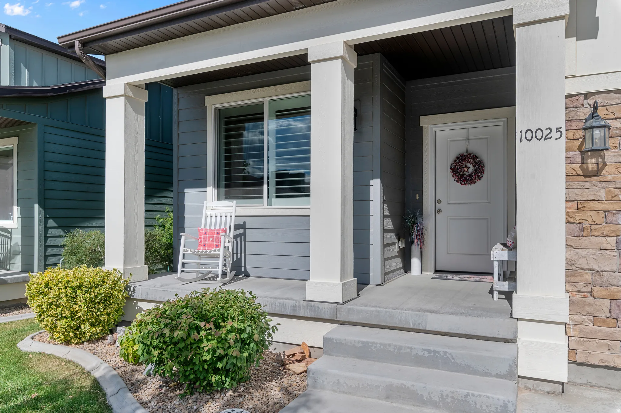 Covered front porch featuring a white entry door, stone veneer accents, gray siding, and a prominent address number