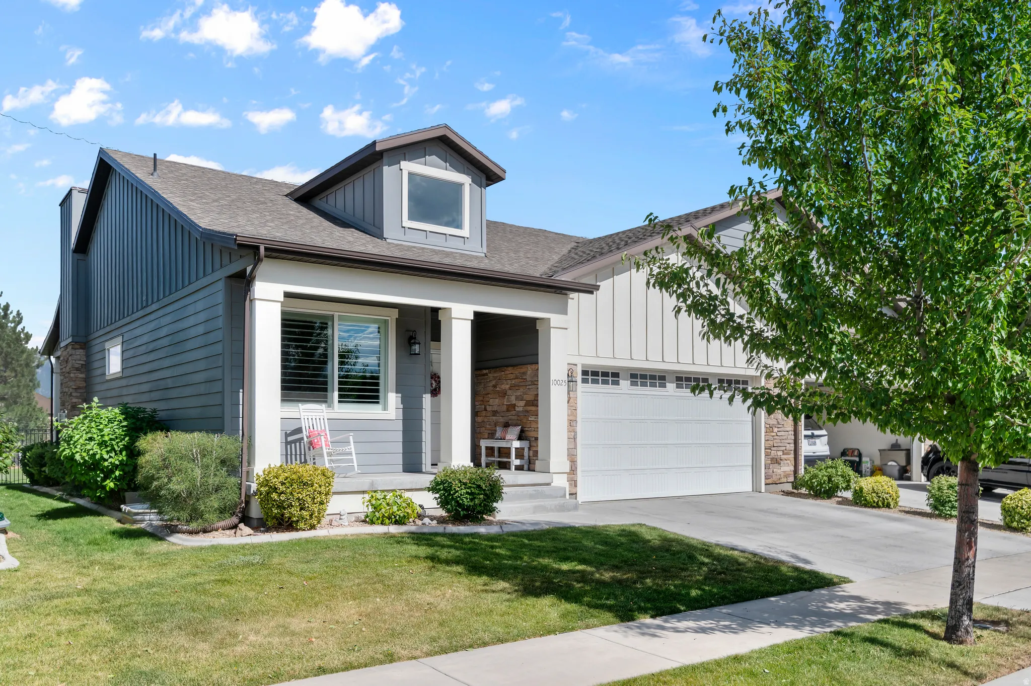 HOME EXTERIOR - FRONT Contemporary home exterior featuring a front porch with stone accents, gray horizontal siding, vertical board and batten siding, a prominent dormer window, and an attached two-car garage