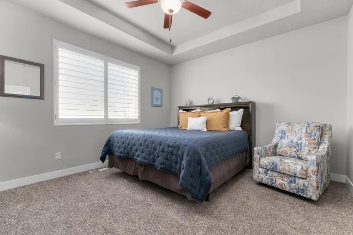 Bedroom featuring a tray ceiling with a ceiling fan, neutral carpet flooring, and a window with plantation shutters