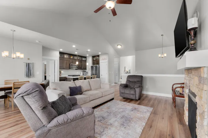 Living area featuring wood-finish flooring, a stone-faced fireplace with a white mantel, and a vaulted ceiling with a ceiling fan
