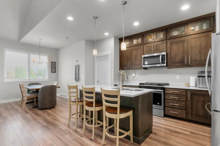 Open-concept kitchen featuring wood-finish flooring, dark wood cabinetry with some upper glass fronts, a central island with a white countertop and seating, stainless steel appliances, and recessed lighting