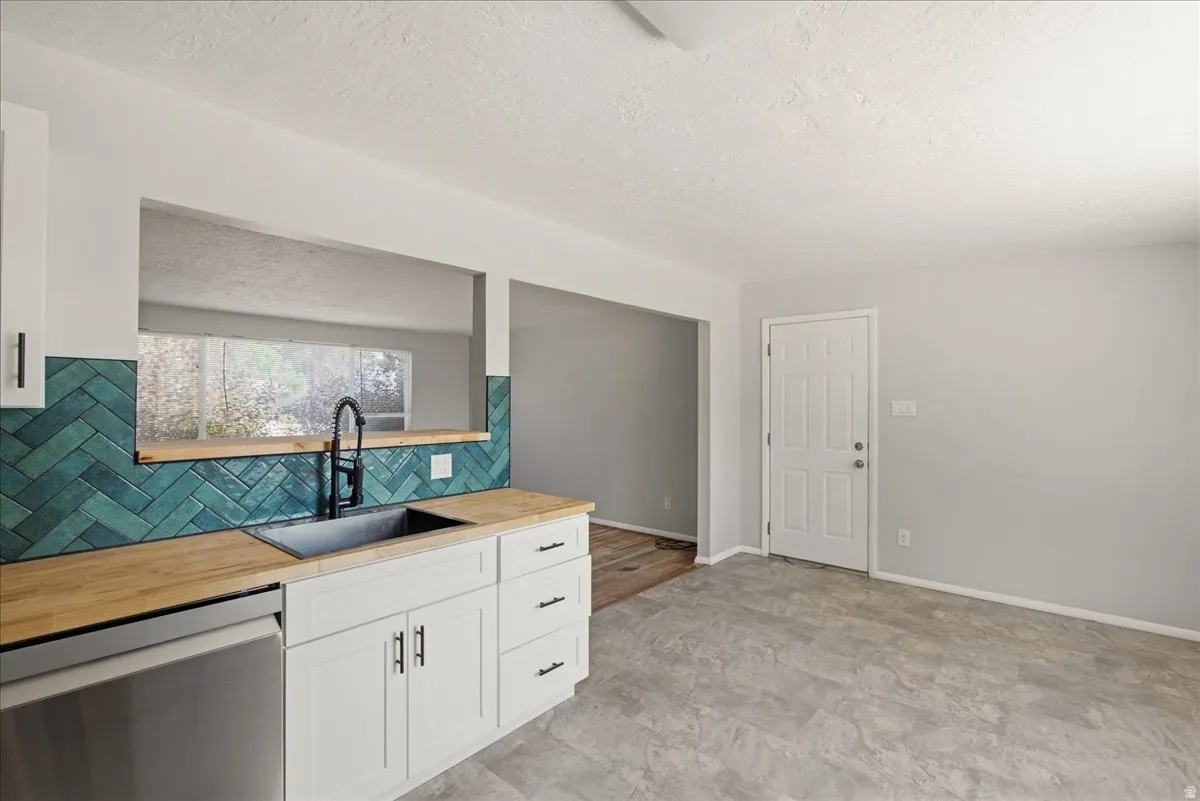 Kitchen featuring white cabinetry with dark hardware, butcher block countertops, a black sink with a black pull-down faucet, a teal herringbone pattern tile backsplash, and stainless steel dishwasher