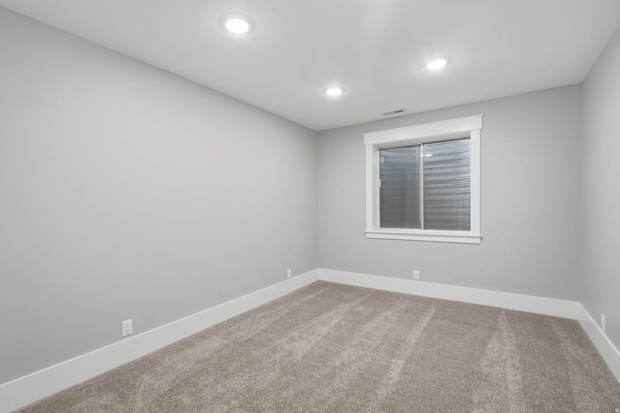Spacious room featuring neutral grey wall paint, plush carpet flooring, recessed ceiling lighting, a white-framed window, and white baseboard trim