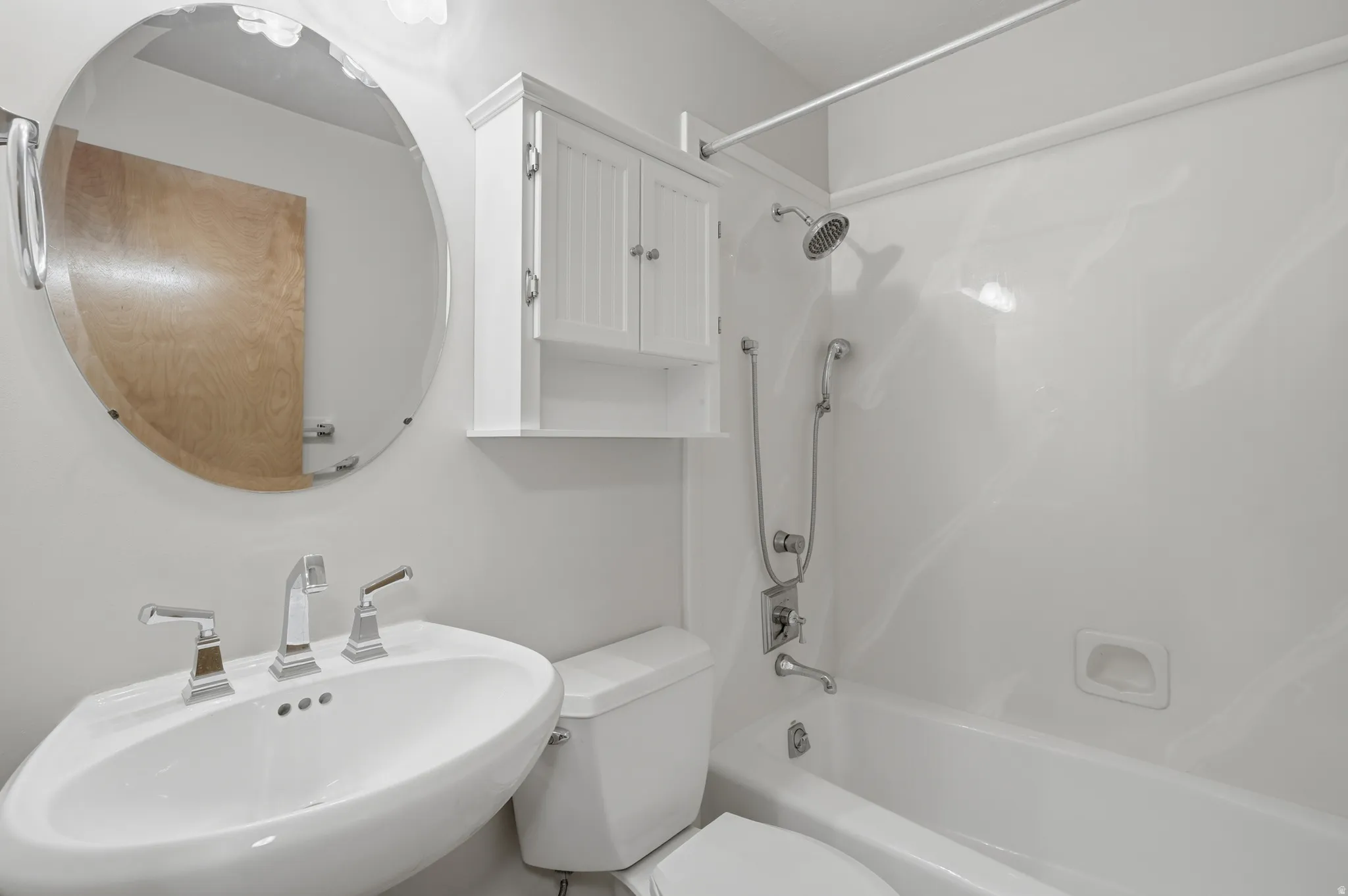Bathroom featuring a pedestal sink with chrome fixtures, a circular frameless mirror, and a built-in wall cabinet