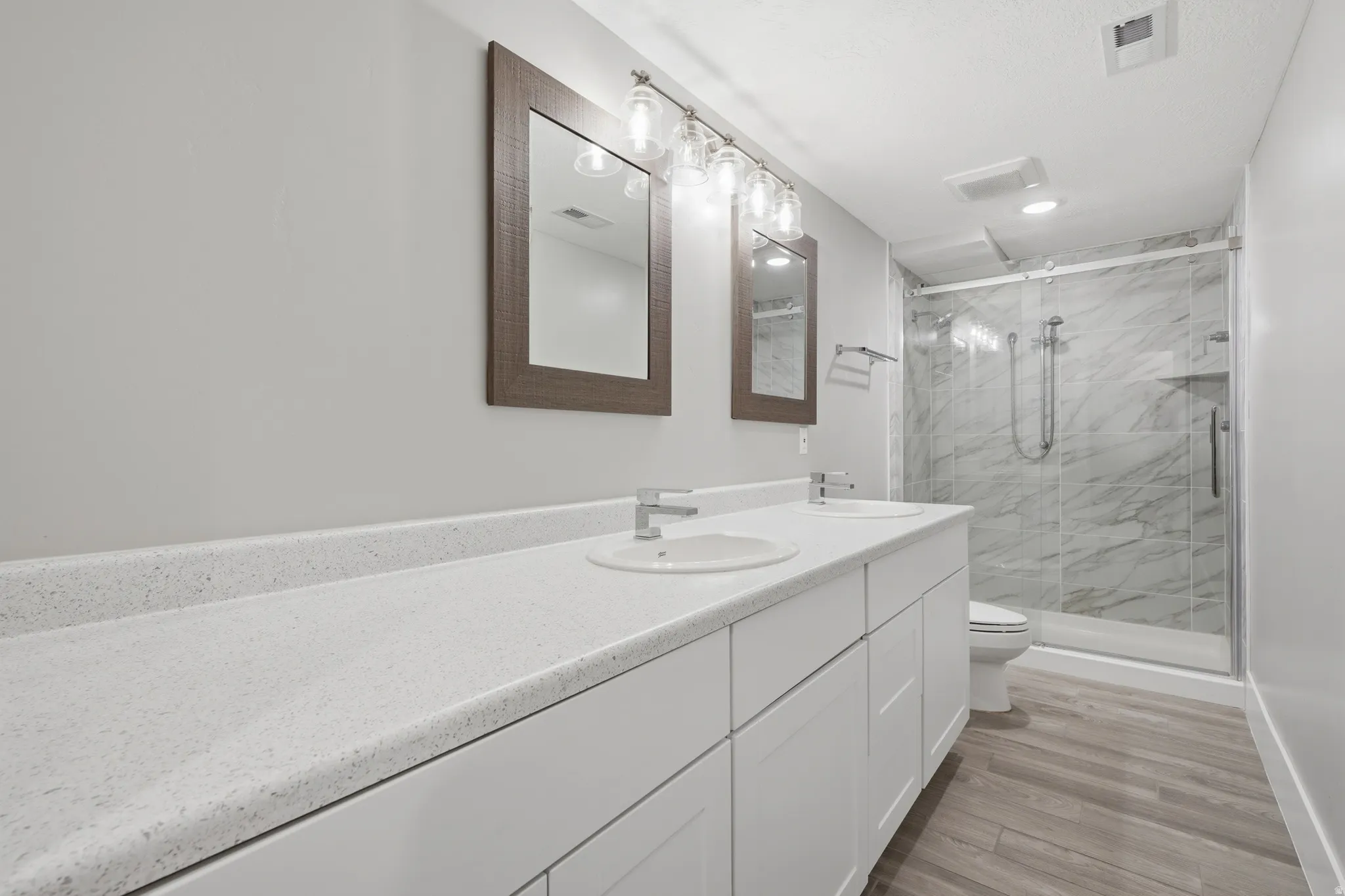 Dual vanity with white cabinetry and light-toned countertops, featuring a tiled shower with a clear glass enclosure, and wood-finish flooring