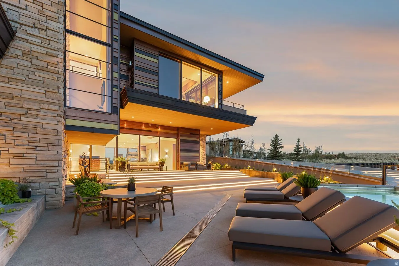 Stone and wood exterior with expansive windows, multi-level patio with integrated lighting, and a swimming pool
