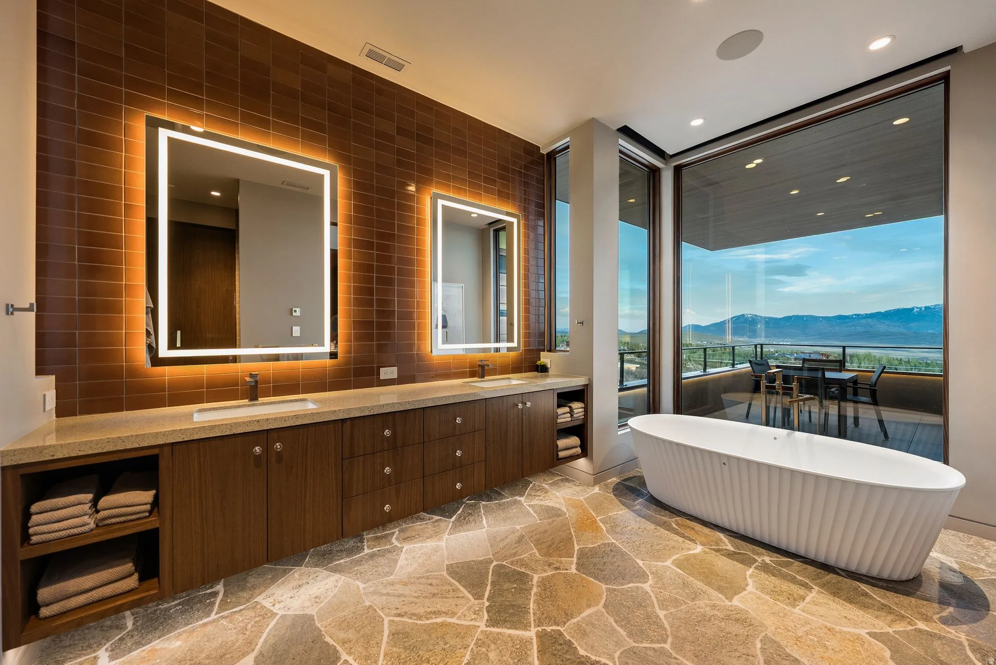 Freestanding soaking tub, stone-finish flooring, full-height windows, dual vanity with undermount sinks, and back-lit mirrors
