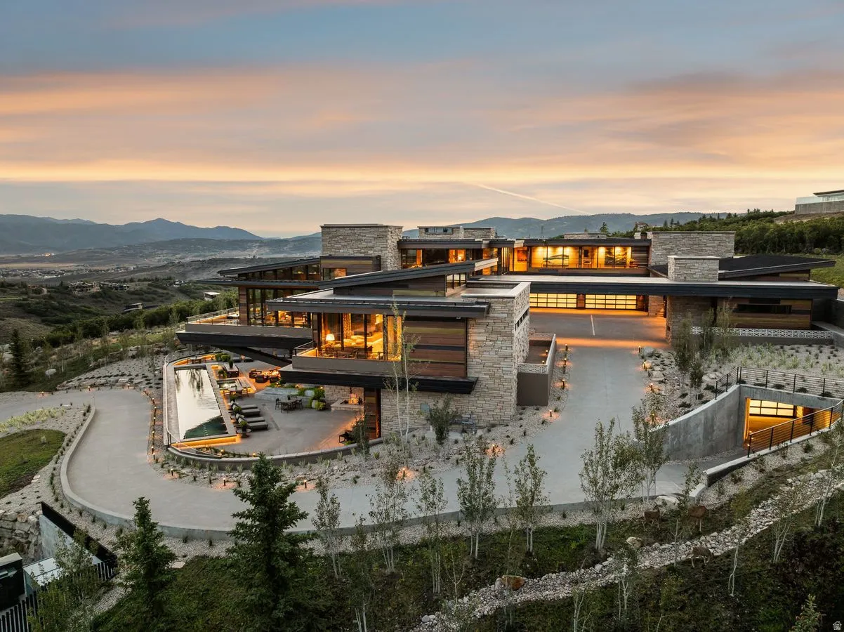Expansive modern residence featuring stone and wood-finish exterior, large windows, multiple balconies, a long driveway, and an outdoor pool
