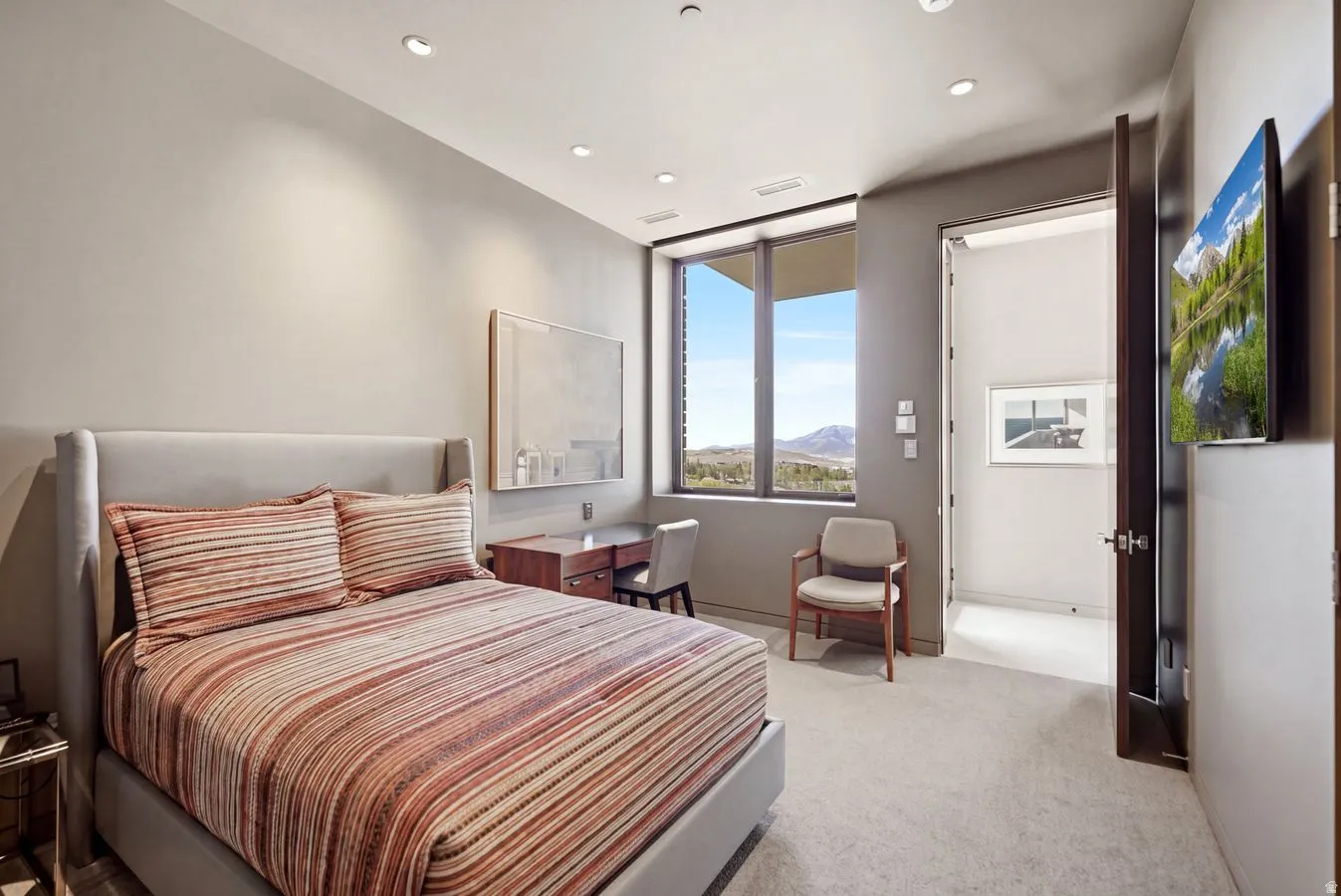 Recessed lighting, wood-finish desk with integrated storage, large window with expansive outdoor scenery, wall-mounted television, and light-toned carpeting