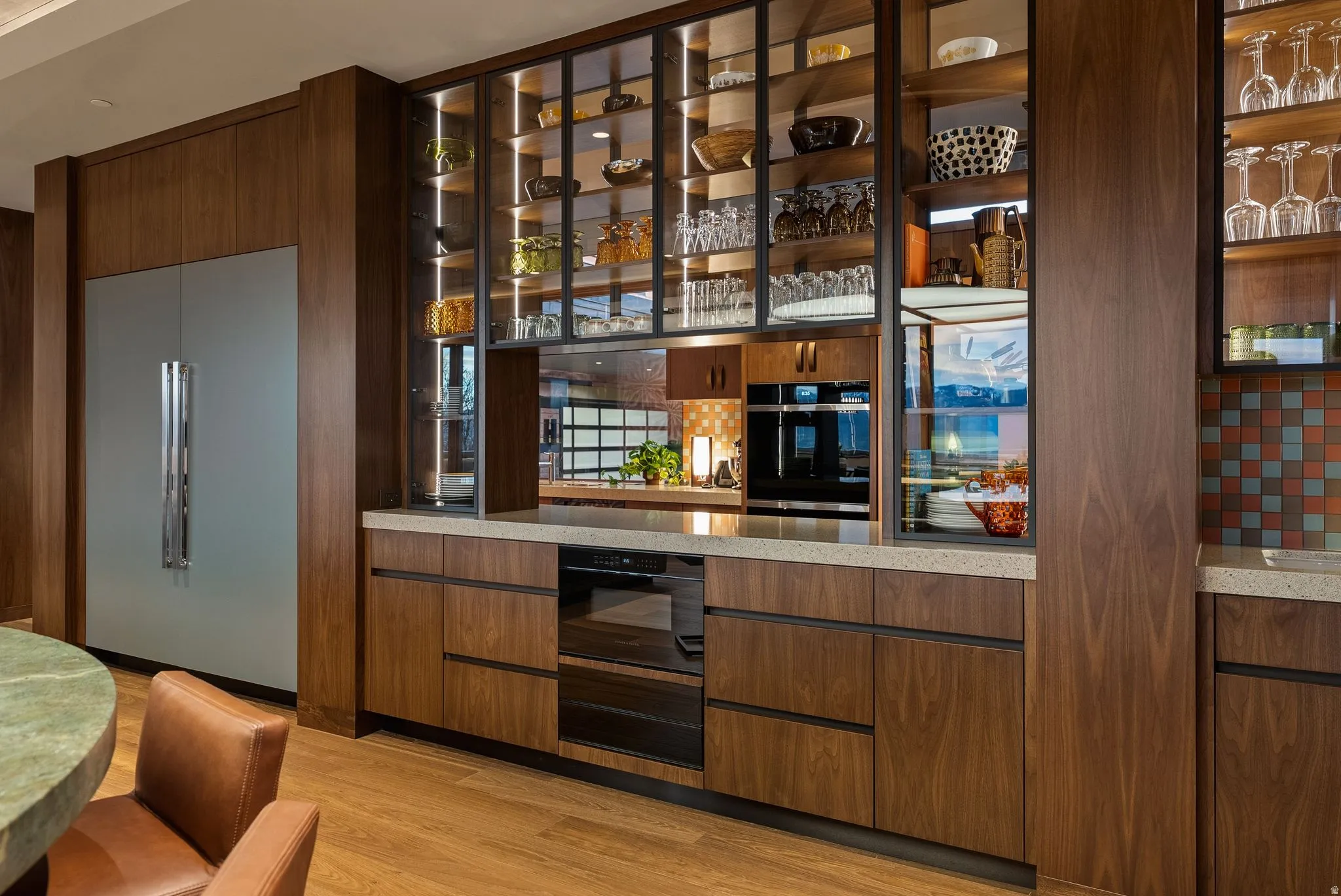 Integrated stainless steel refrigerator, custom wood cabinetry, extensive glass-front display shelving with interior lighting, built-in wall oven, and light-toned countertops