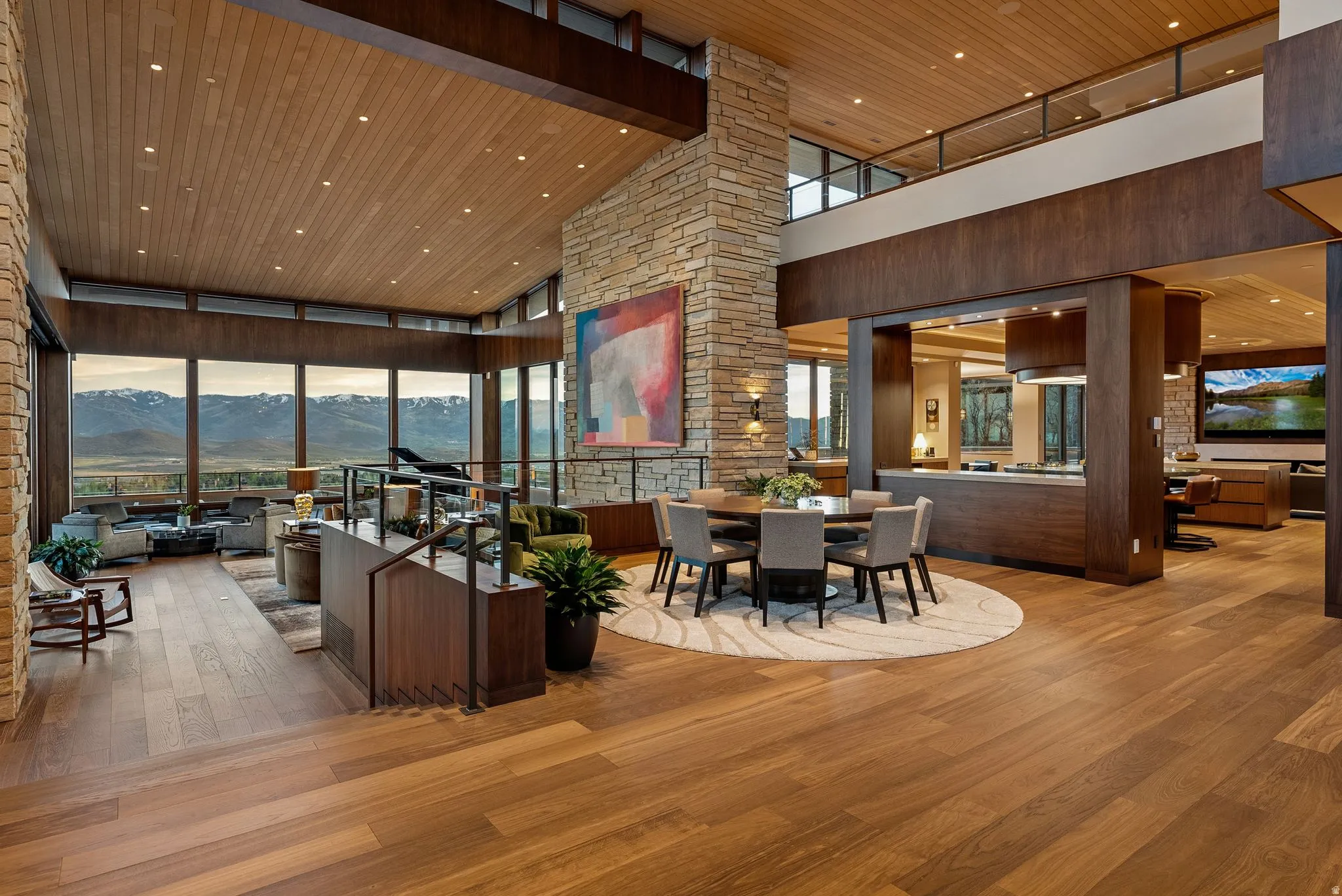 Expansive open-concept living area featuring wood-finish flooring, a stone-clad pillar, a vaulted wood-paneled ceiling, and a dining area with a circular table