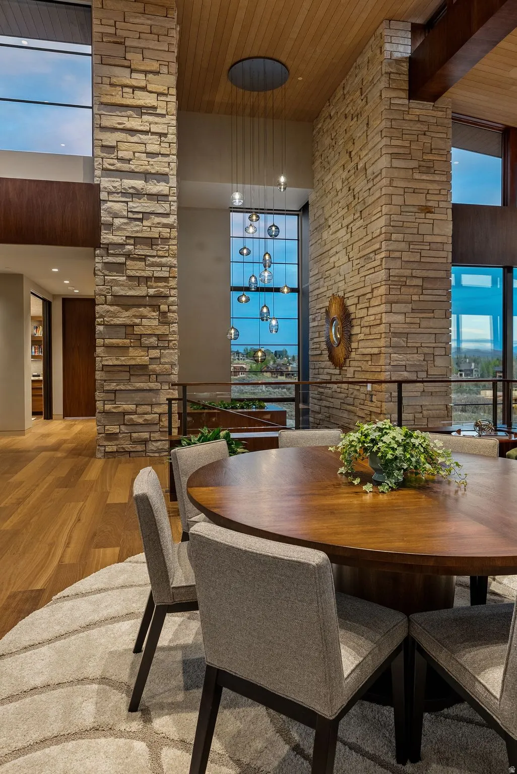 Soaring ceiling with wood-finish paneling, stone-clad architectural columns, floor-to-ceiling windows, and hardwood flooring