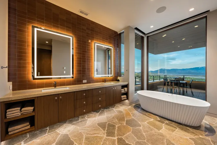 Freestanding soaking tub, stone-finish flooring, full-height windows, dual vanity with undermount sinks, and back-lit mirrors