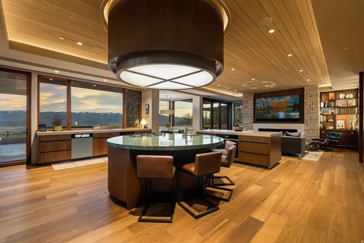 Hardwood flooring throughout, a large circular island with an integrated cooktop, a stone fireplace, built-in shelving, and extensive windows