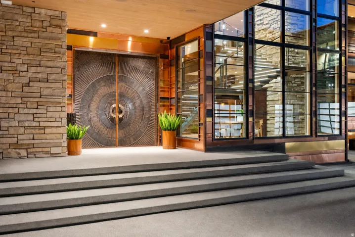 Textured stone facade, grand double entry doors with intricate circular detailing, multi-level exterior glass panels, metal-framed windows, and broad concrete entry steps