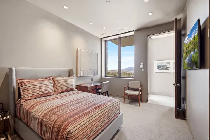 Recessed lighting, wood-finish desk with integrated storage, large window with expansive outdoor scenery, wall-mounted television, and light-toned carpeting
