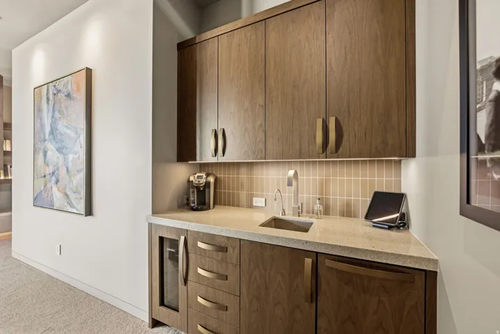 Integrated wet bar featuring wood-finish cabinetry, a stone-finish countertop, under-cabinet lighting, a tiled backsplash, and a stainless steel sink with a gooseneck faucet