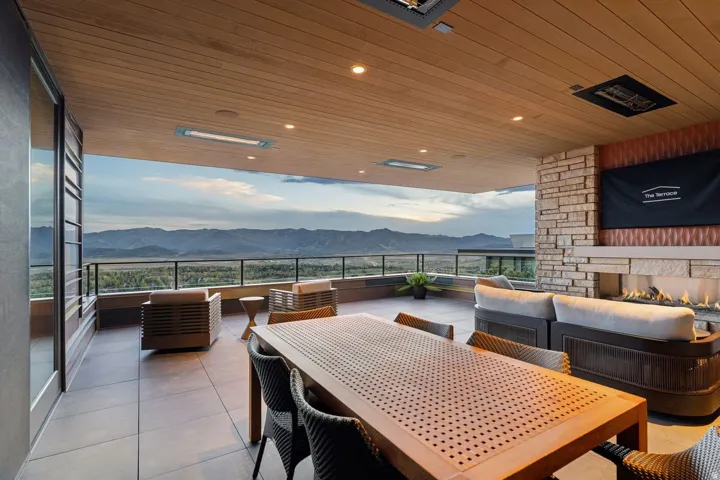 Expansive covered patio featuring a wood-finish ceiling, mounted outdoor heaters, a stacked stone fireplace, and a glass-paneled railing