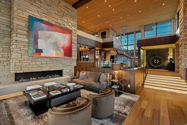 Two-story stone-clad fireplace with linear hearth, wood-finish flooring, integrated staircase with metal railing, and high ceilings with wood paneling