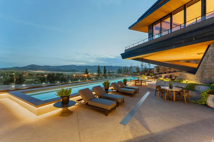 Infinity-edge pool with perimeter lighting, expansive patio space, elevated architectural design, extensive window features, and surrounding landscape