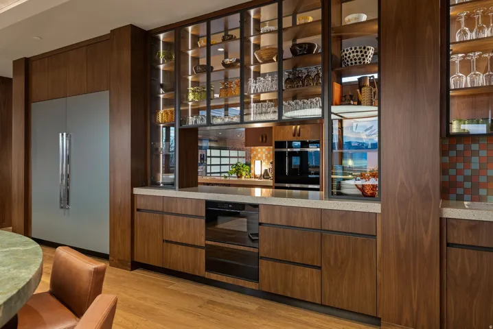 Integrated stainless steel refrigerator, custom wood cabinetry, extensive glass-front display shelving with interior lighting, built-in wall oven, and light-toned countertops