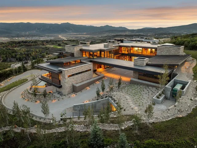 Expansive contemporary residence featuring stone and wood exterior elements, multiple levels of outdoor decking, a winding paved driveway, and mature landscaping