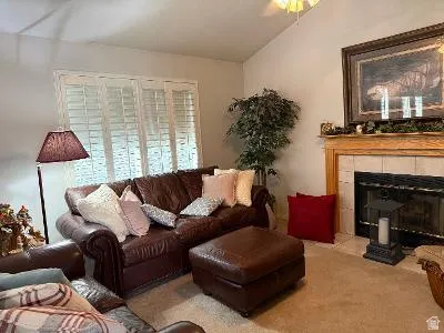 Living area featuring a fireplace with a tile surround and wood mantel, vaulted ceiling, ceiling fan, and window with plantation shutters