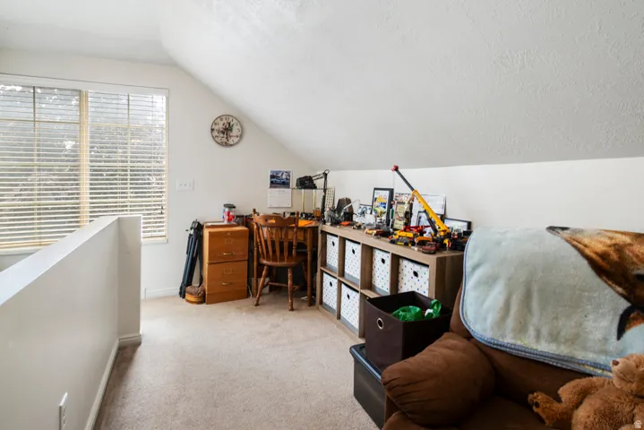 Carpeted loft area featuring a large window with horizontal blinds, sloped ceiling, and a built-in shelving unit