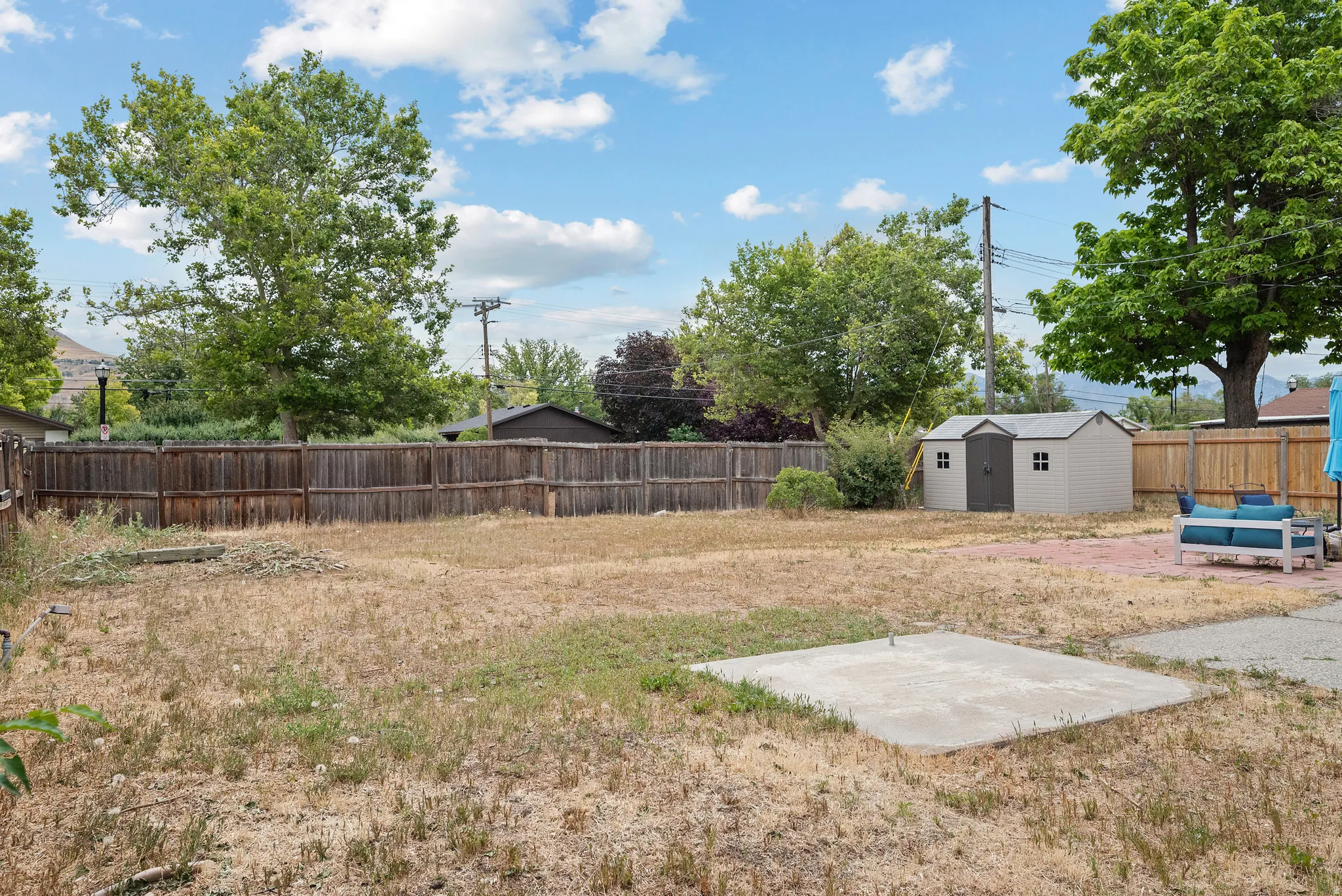 Expansive outdoor space featuring a perimeter wood fence, a detached storage shed, a concrete patio area, and mature trees