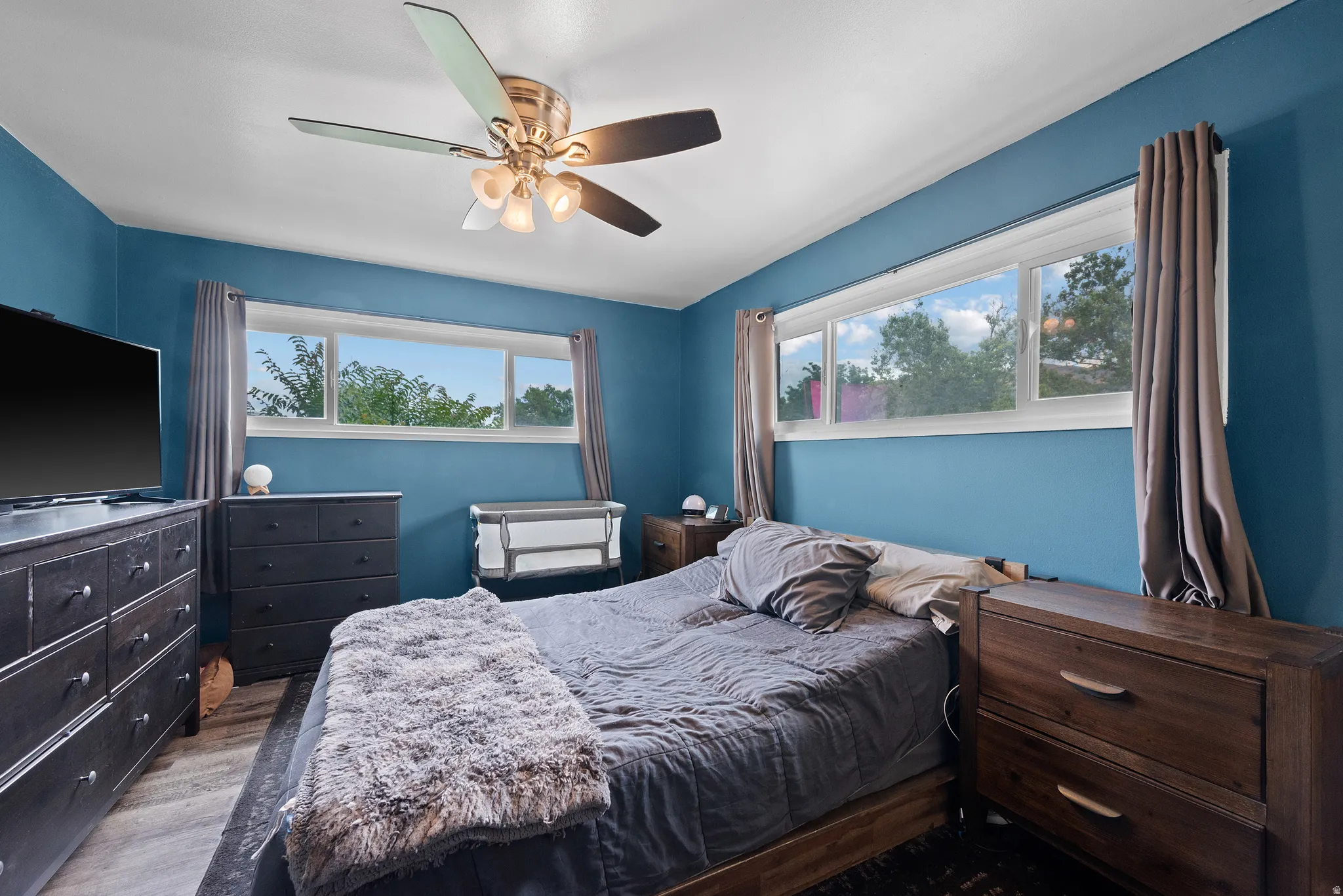 Bedroom featuring wood-finish flooring, two large horizontal windows, a ceiling fan with integrated lighting, and painted walls