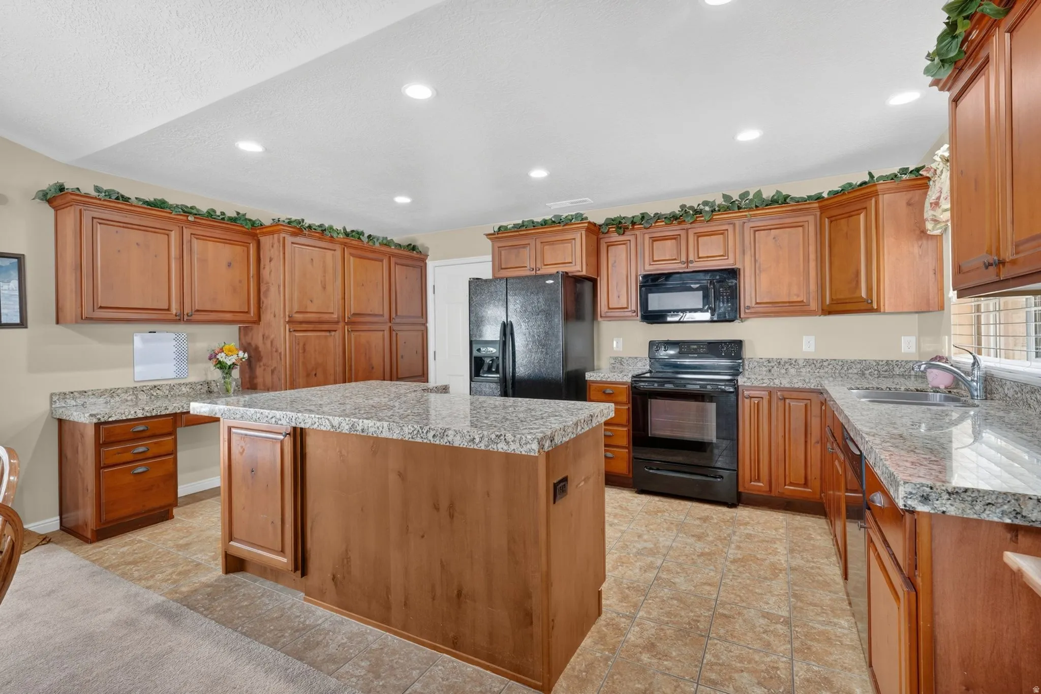 Spacious kitchen featuring an expansive central island, granite countertops, wood cabinetry, built-in black appliances, and tile flooring