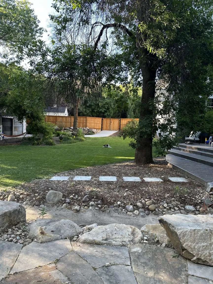 Expansive backyard featuring a natural stone patio, a large grass lawn, mature trees, a wood privacy fence, and concrete stepping stones