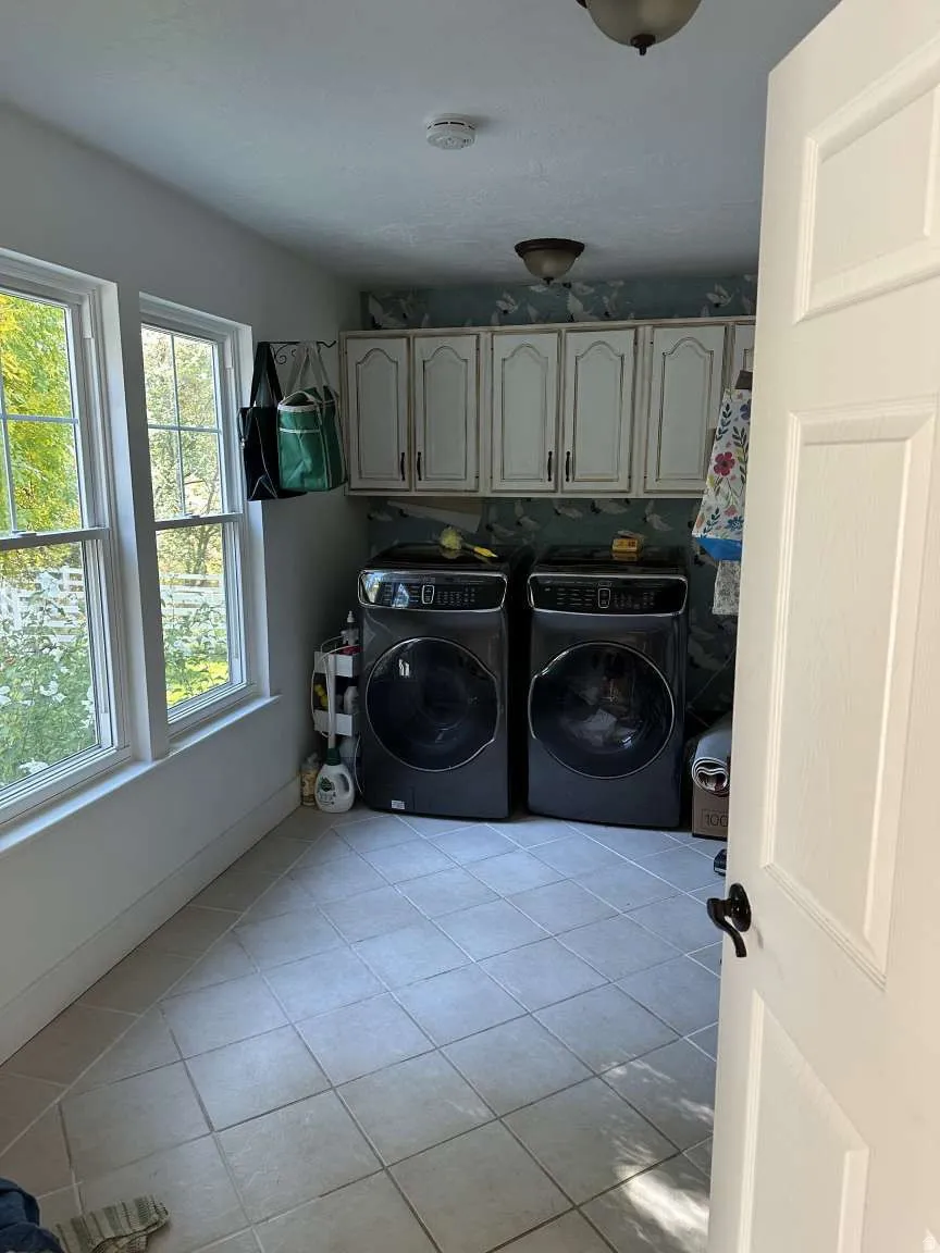 Laundry space featuring light-toned tile flooring, two large windows, white cabinetry with dark hardware, and a ceiling-mounted light fixture