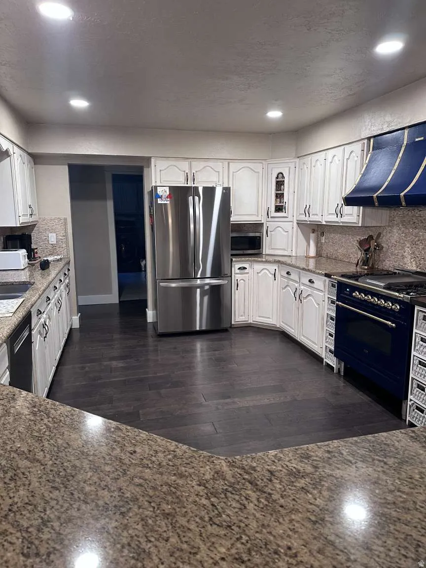 Kitchen featuring granite-style countertops, dark wood-finish flooring, white cabinetry with decorative trim, a stainless steel refrigerator, and a blue range with a matching custom hood