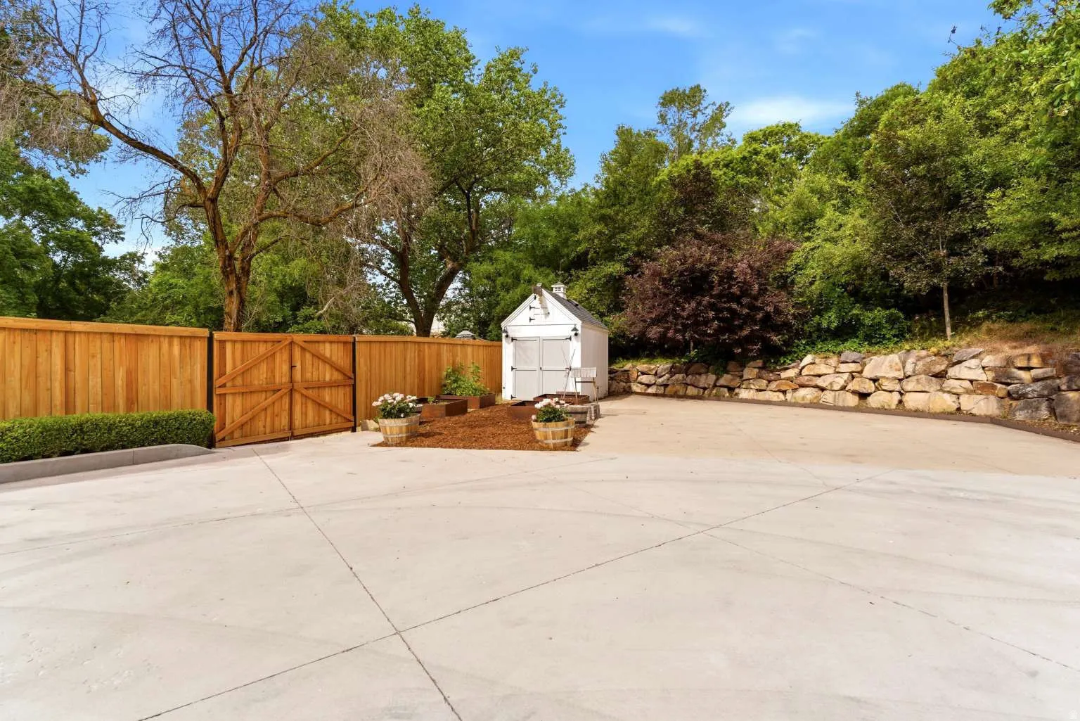 Spacious concrete pad featuring a privacy fence with double gates, a white storage shed, and a natural stone retaining wall