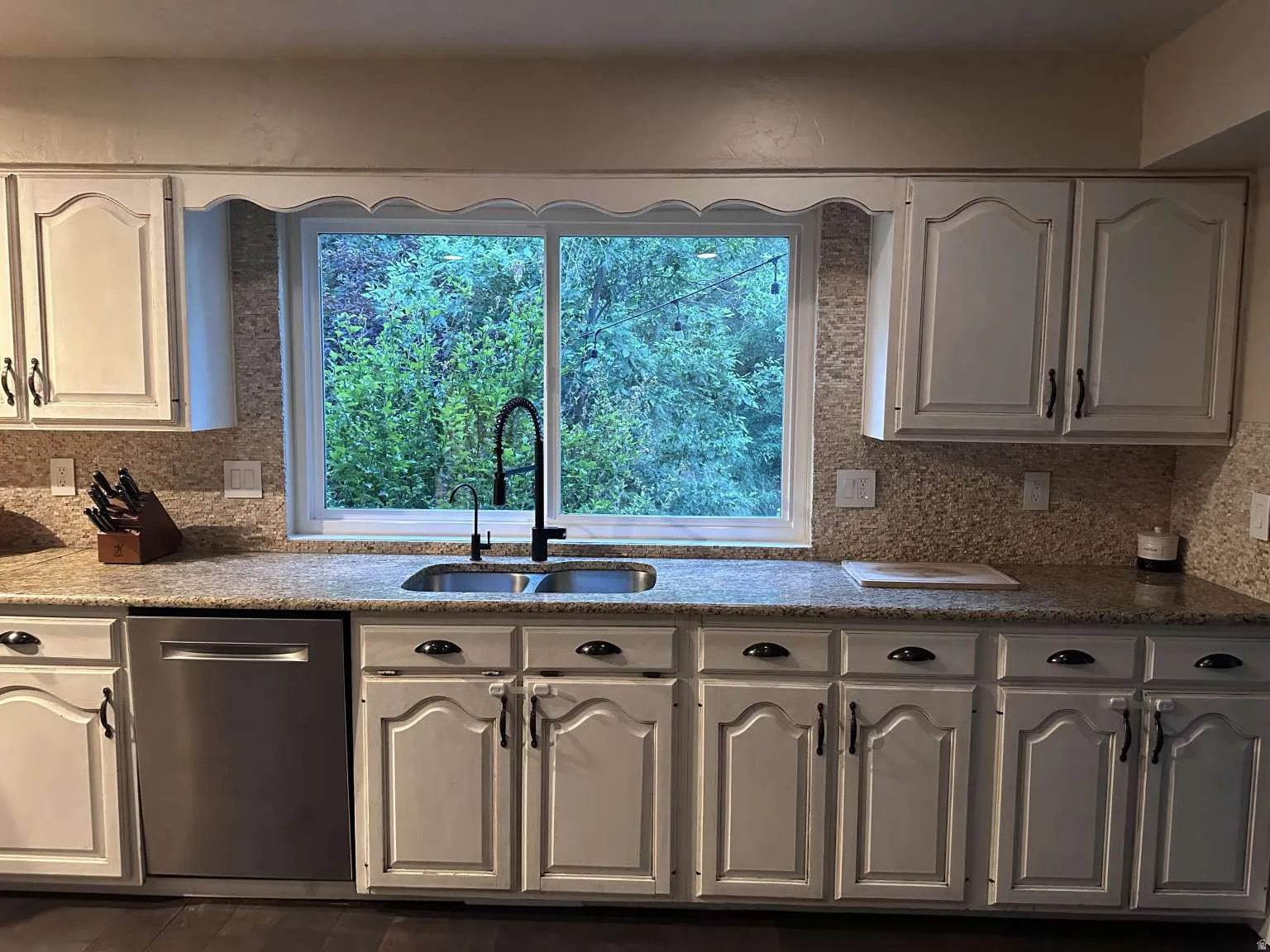 Kitchen featuring light-finish cabinetry with dark hardware, a double basin sink with a black gooseneck faucet, light granite-finish countertops, and a patterned tile backsplash