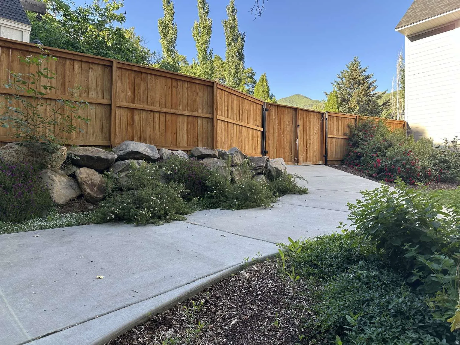 Private side yard featuring a wood privacy fence, concrete pathway, and a rock retaining wall with established plantings