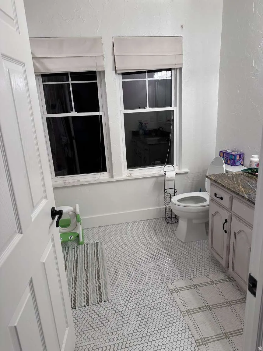 Bathroom featuring white hexagonal tile flooring, dual windows with Roman shades, a vanity with a stone countertop, and a white paneled door with black hardware