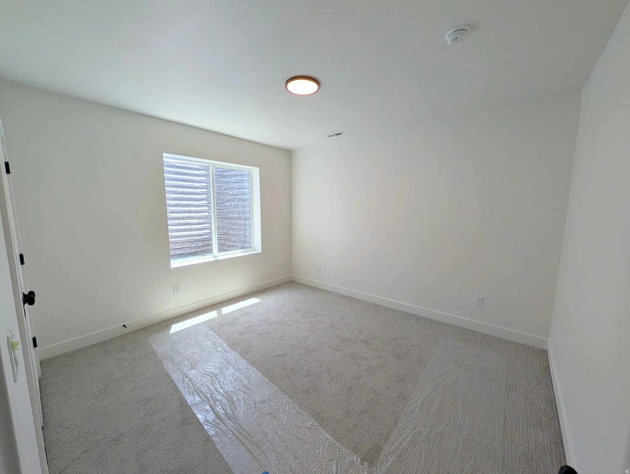 Neutral-toned room featuring a single window with horizontal blinds, light-colored carpeting, a ceiling-mounted light fixture, and white painted walls