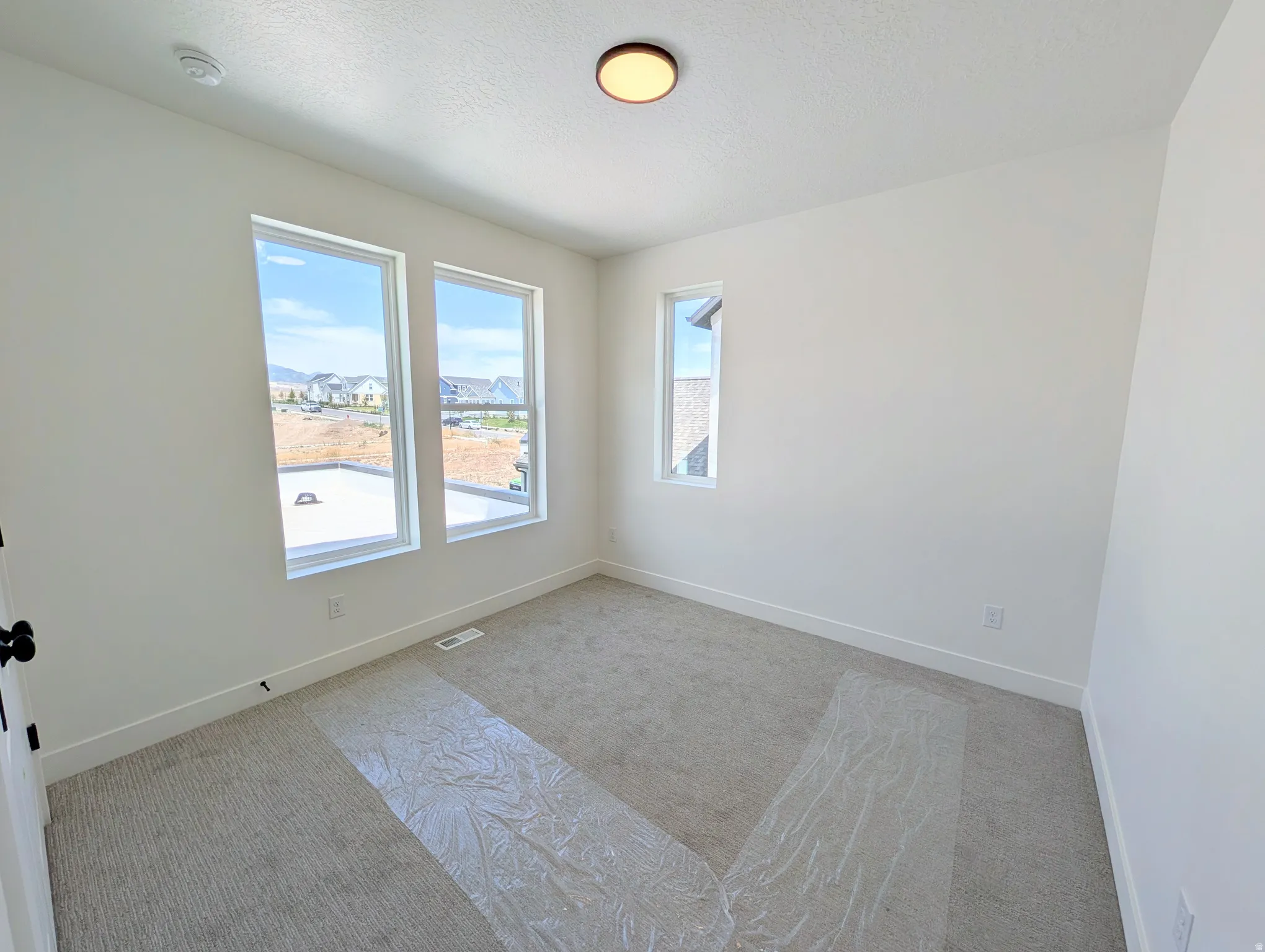 Bright room featuring three windows, a ceiling-mounted light fixture, light gray carpeting, crisp white walls, and white baseboards