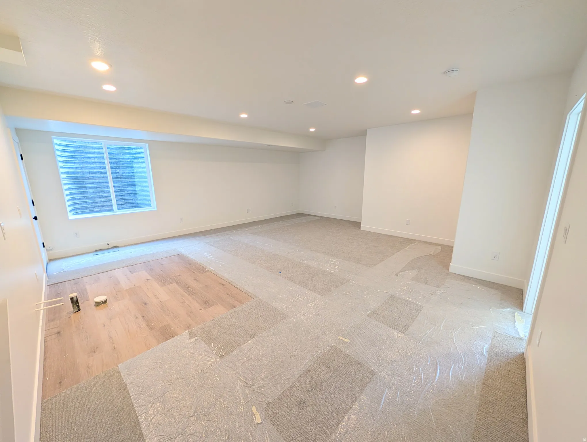 Spacious room featuring recessed lighting, a large egress window with white blinds, white baseboards, and a combination of unfinished wood-finish flooring and protected carpet