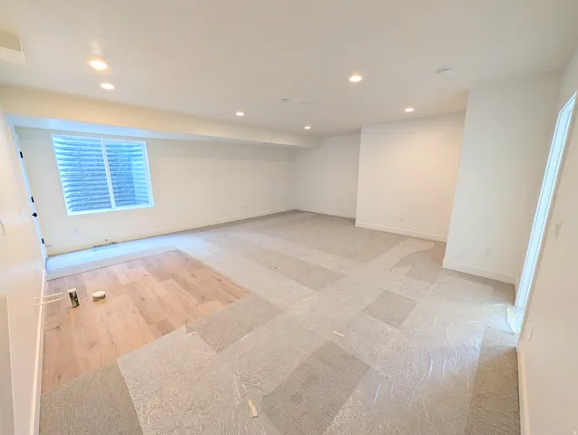 Spacious room featuring recessed lighting, a large egress window with white blinds, white baseboards, and a combination of unfinished wood-finish flooring and protected carpet