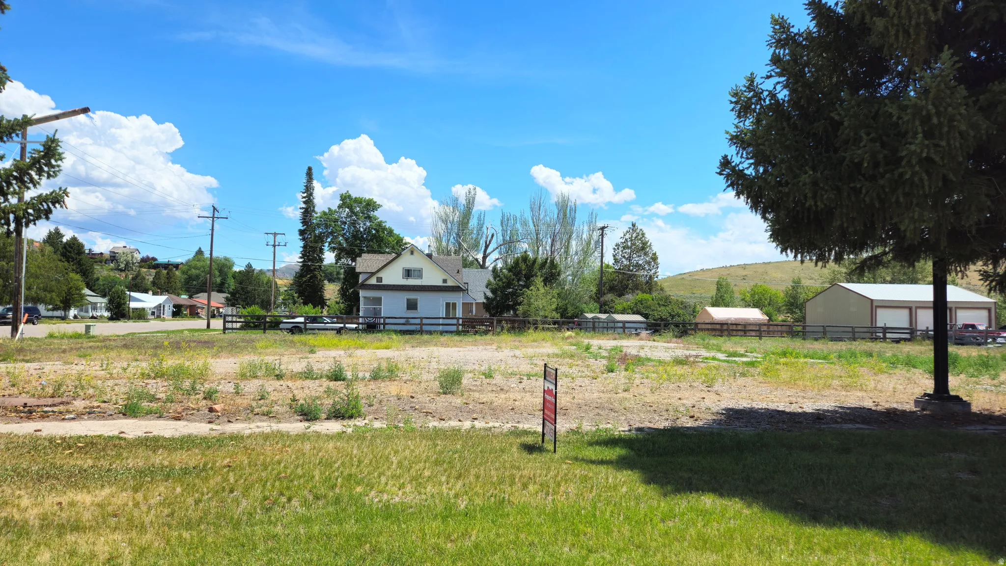 Expansive land parcel with partial grass coverage, featuring distant residential structures, utility poles, and mature trees