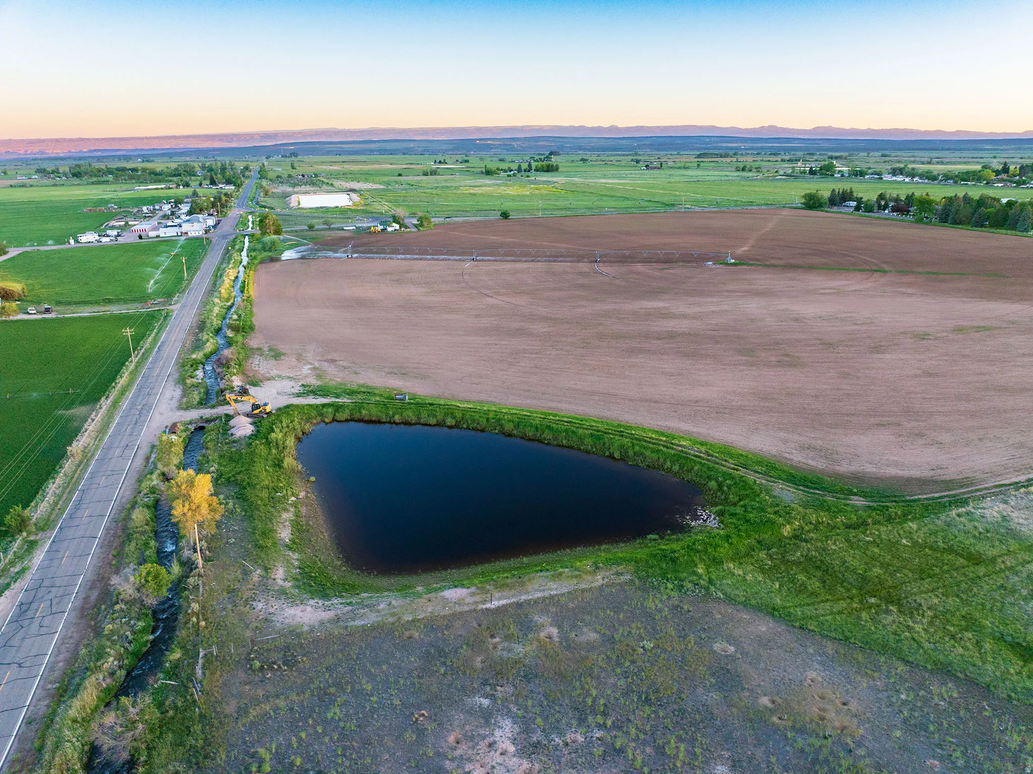 Expansive agricultural land featuring a natural pond, paved road frontage, and irrigation ditches