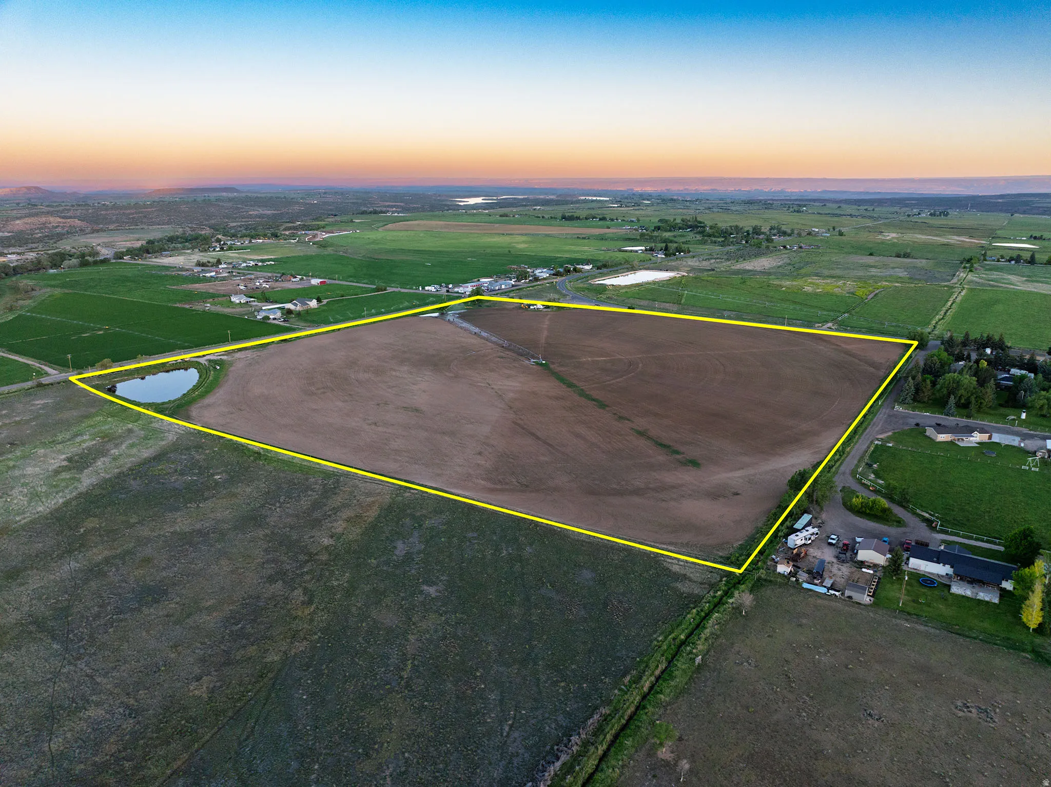 Expansive agricultural land featuring a natural pond, surrounding green fields, and distant residential structures