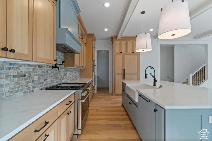 Expansive kitchen featuring wood-finish flooring, light wood cabinetry, stone-finish countertops, a stone tile backsplash, and an island with an integrated farmhouse sink