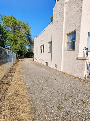 Stucco exterior with multiple windows, concrete walkway, chain link fencing, and mature trees