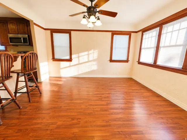 Open-concept living area featuring wood-finish flooring, multiple windows with blinds, a ceiling fan with integrated lighting, and a partial kitchen pass-through with built-in cabinetry and microwave